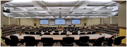 Large classroom, well lit, with tables and chairs in rows, three large video monitors at the front of the room.