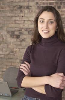 Young, ethnic woman with long, dark hair, with a slight smile, her arms crossed, she's wearing a long-sleeved maroon turtleneck, and standing in front of a desk with a computer against an aged brick wall.